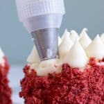 A piping bag applies white frosting onto the top of a red velvet cake with red crumbs on the sides.