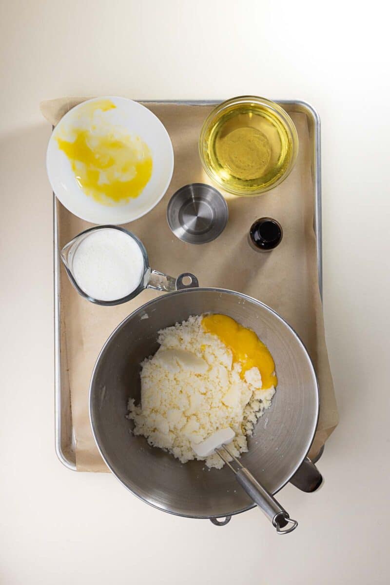 A mixing bowl with sugar, butter, and egg yolks sits on a tray; nearby are a cup of milk, a bowl with more egg yolks, a cup of oil, a small pitcher, and a bottle of vanilla extract.