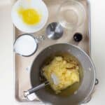 A metal mixing bowl with creamed butter and sugar, surrounded by a cup of milk, a bowl with eggs, a measuring cup, a small bowl of vanilla, and an empty glass bowl on a baking tray.