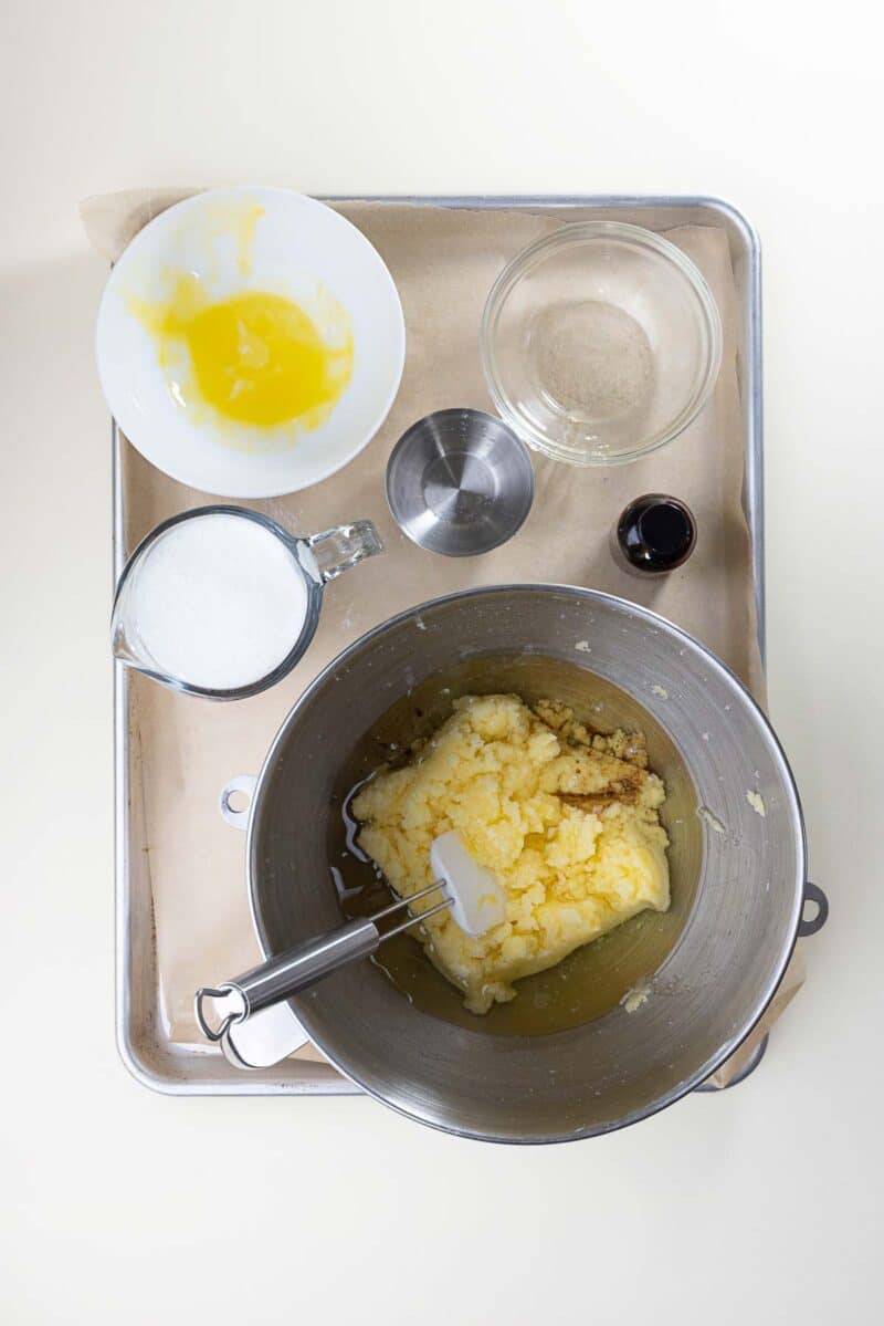 A metal mixing bowl with creamed butter and sugar, surrounded by a cup of milk, a bowl with eggs, a measuring cup, a small bowl of vanilla, and an empty glass bowl on a baking tray.