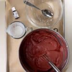 A mixing bowl with red cake batter, a whisk, an empty glass bowl with a spoon, a measuring cup with liquid, and a bottle of food coloring on a parchment-lined tray.