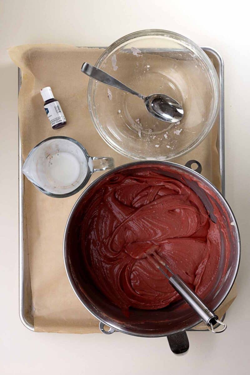 A mixing bowl with red cake batter, a whisk, an empty glass bowl with a spoon, a measuring cup with liquid, and a bottle of food coloring on a parchment-lined tray.