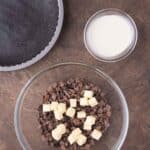A glass bowl with chopped chocolate and butter cubes, a bowl of cream, a chocolate crust in a pan, and a small bowl of berries on a wooden surface.