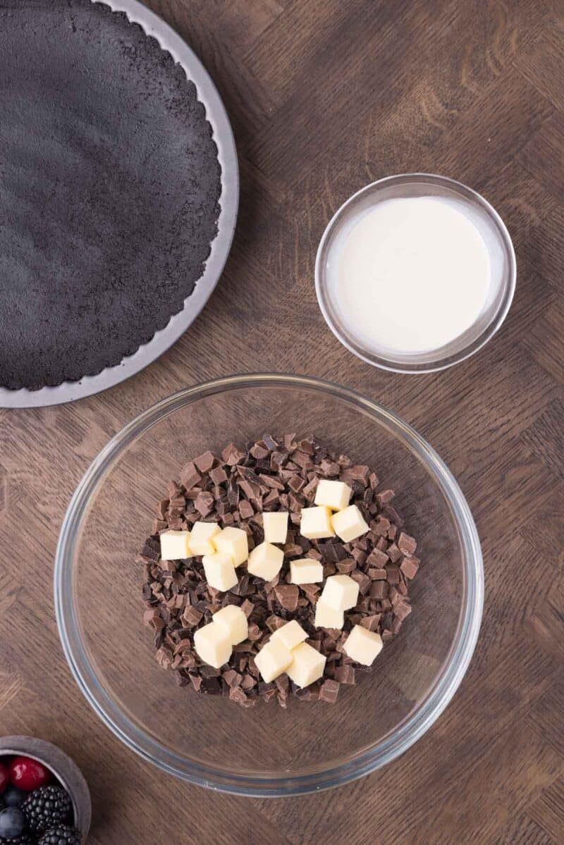A glass bowl with chopped chocolate and butter cubes, a bowl of cream, a chocolate crust in a pan, and a small bowl of berries on a wooden surface.