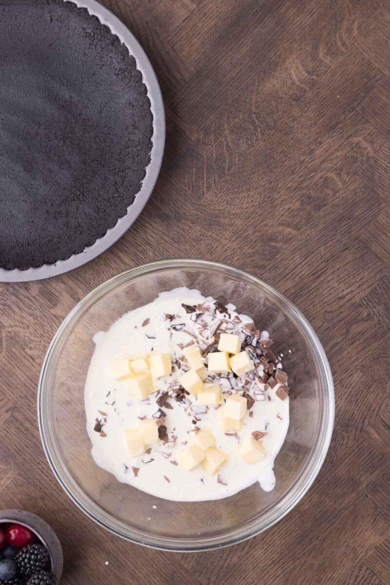 A glass bowl with cream, chocolate pieces, and butter cubes on a wooden surface next to an empty chocolate pie crust and a small bowl of berries.