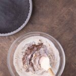 A hand stirs chocolate into a creamy mixture in a glass bowl with a spatula; an empty chocolate crust and a bowl of berries sit nearby on a wooden surface.