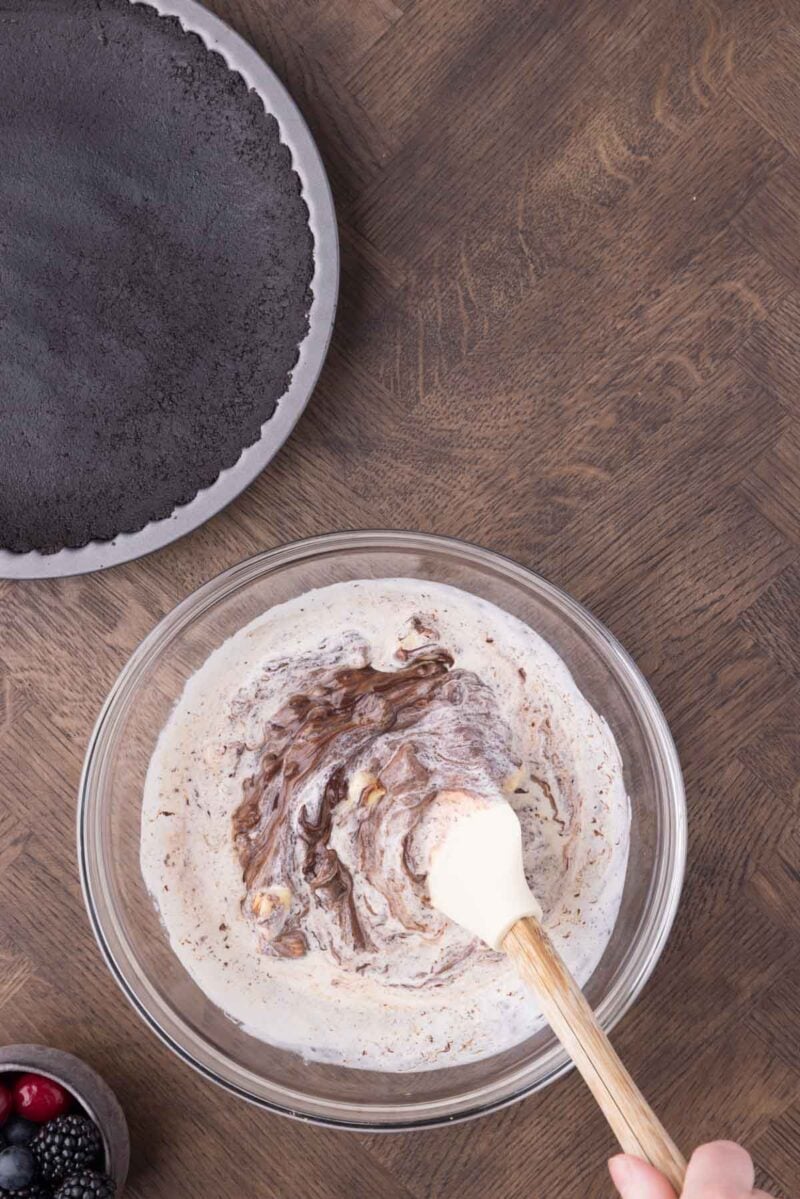 A hand stirs chocolate into a creamy mixture in a glass bowl with a spatula; an empty chocolate crust and a bowl of berries sit nearby on a wooden surface.