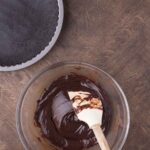 A hand holds a spatula mixing chocolate filling in a glass bowl next to a prepared chocolate pie crust and a small bowl of berries on a wooden surface.