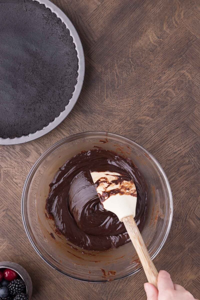 A hand holds a spatula mixing chocolate filling in a glass bowl next to a prepared chocolate pie crust and a small bowl of berries on a wooden surface.