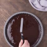 A hand uses an offset spatula to spread chocolate ganache over a tart crust in a round metal pan on a wooden table.