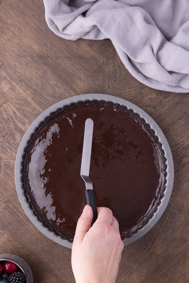A hand uses an offset spatula to spread chocolate ganache over a tart crust in a round metal pan on a wooden table.