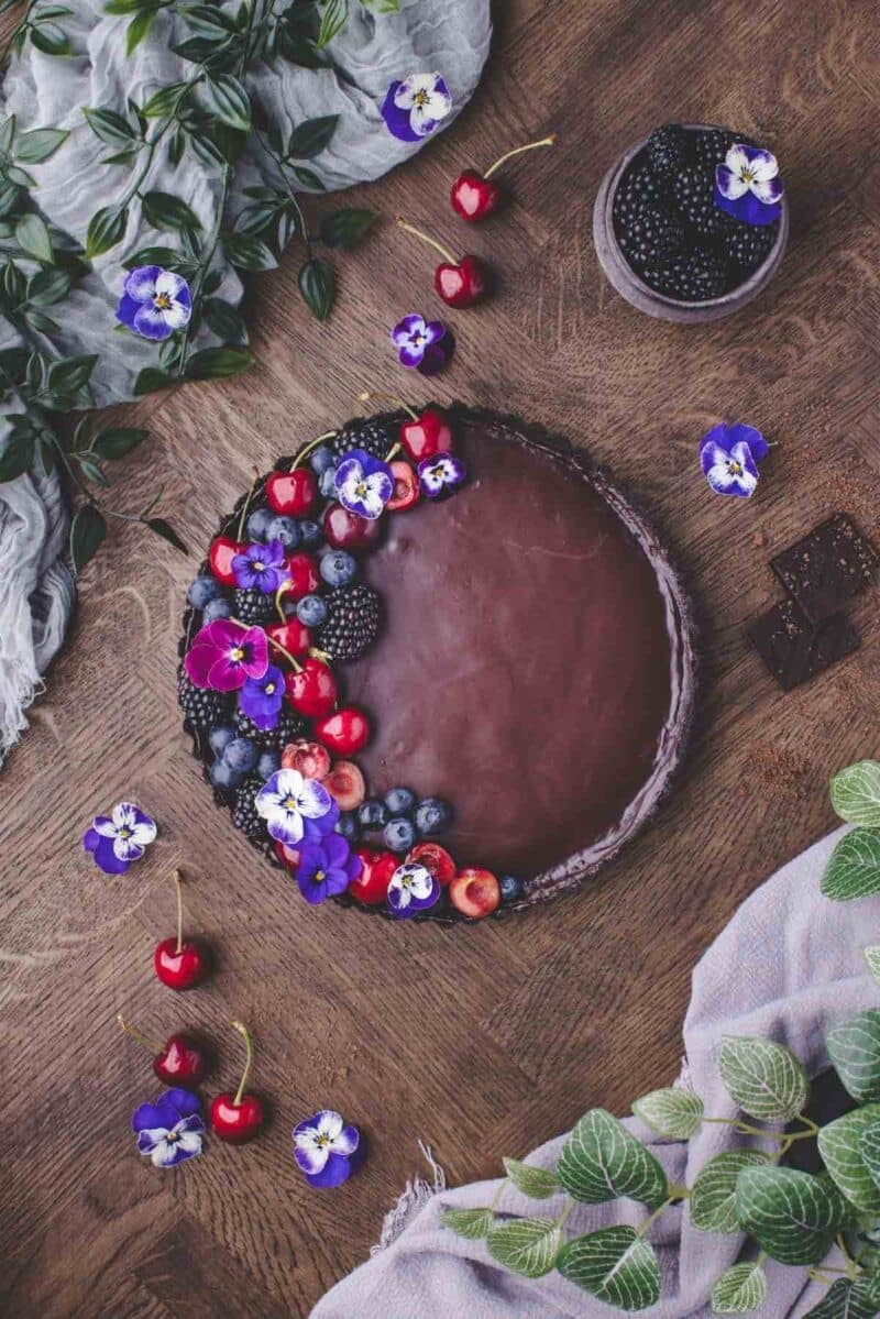 A chocolate cake topped with assorted berries, cherries, and edible purple flowers on a wooden table with green leaves and a bowl of blackberries nearby.