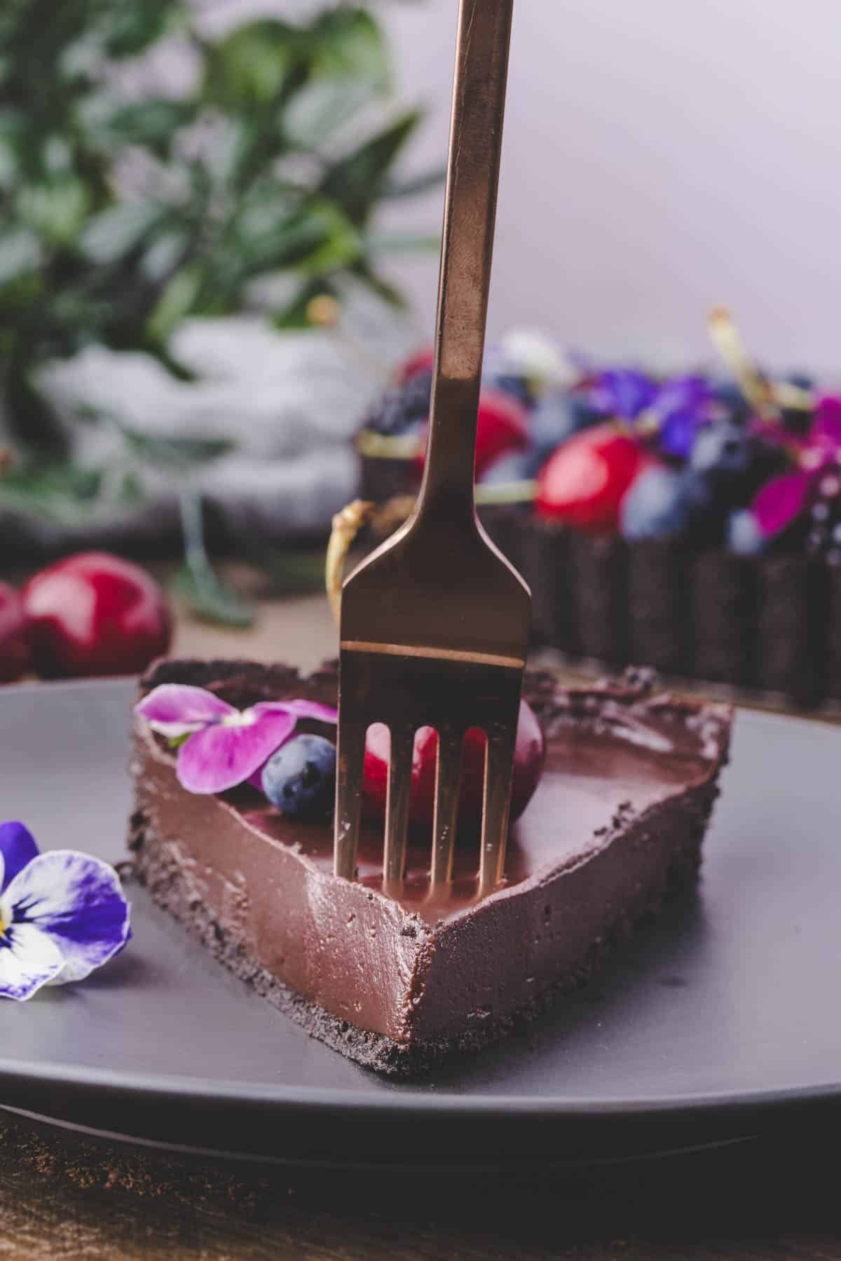 A fork presses into a slice of chocolate tart topped with berries and edible flowers on a gray plate.