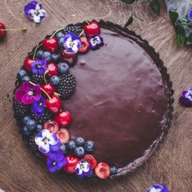 A chocolate tart topped with cherries, blueberries, blackberries, and purple edible flowers sits on a wooden table.
