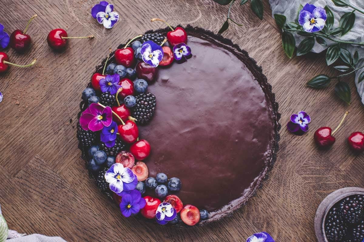 A chocolate tart topped with cherries, blueberries, blackberries, and purple edible flowers sits on a wooden table.