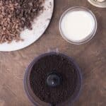 A food processor with crushed cookies, a bowl of cream, a bowl of butter cubes, chopped chocolate on a plate, and a small bowl of berries on a wooden surface.