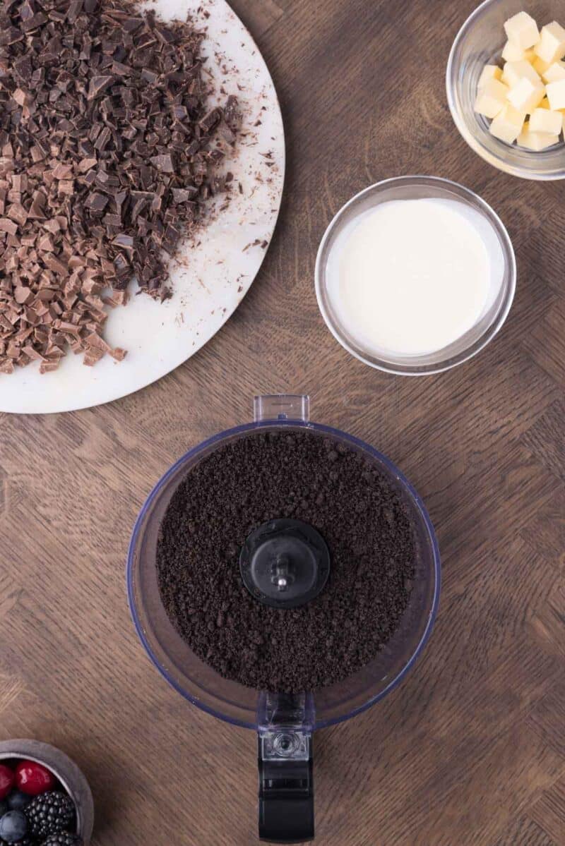 A food processor with crushed cookies, a bowl of cream, a bowl of butter cubes, chopped chocolate on a plate, and a small bowl of berries on a wooden surface.