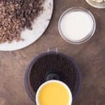 A hand pours melted butter into a food processor with chocolate cookie crumbs; nearby are bowls of cream, butter cubes, chopped chocolate, and mixed berries.