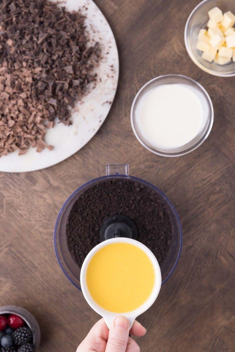 A hand pours melted butter into a food processor with chocolate cookie crumbs; nearby are bowls of cream, butter cubes, chopped chocolate, and mixed berries.