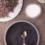 A hand presses crushed chocolate cookie crumbs into a tart pan with a spoon; nearby are chopped chocolate, cream, butter, and a small bowl of berries.