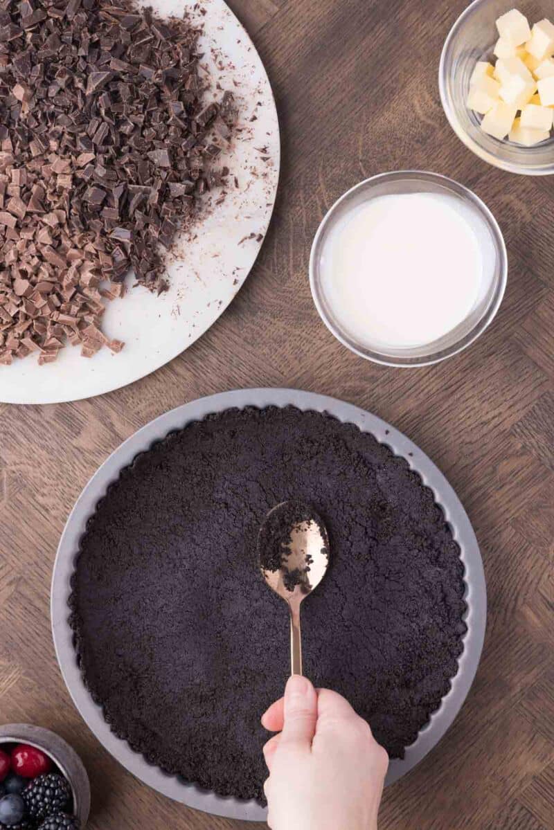 A hand presses crushed chocolate cookie crumbs into a tart pan with a spoon; nearby are chopped chocolate, cream, butter, and a small bowl of berries.