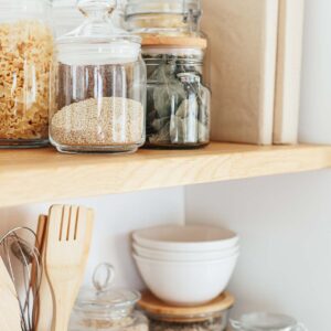 A wooden kitchen shelf holds glass jars of dry goods, white bowls, utensils in a holder, and a stack of beige books or boxes against a white wall.