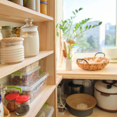 Organized pantry with shelves holding jars and containers of dry goods, a counter with kitchen appliances, a basket of onions, and a potted plant by a sunny window.