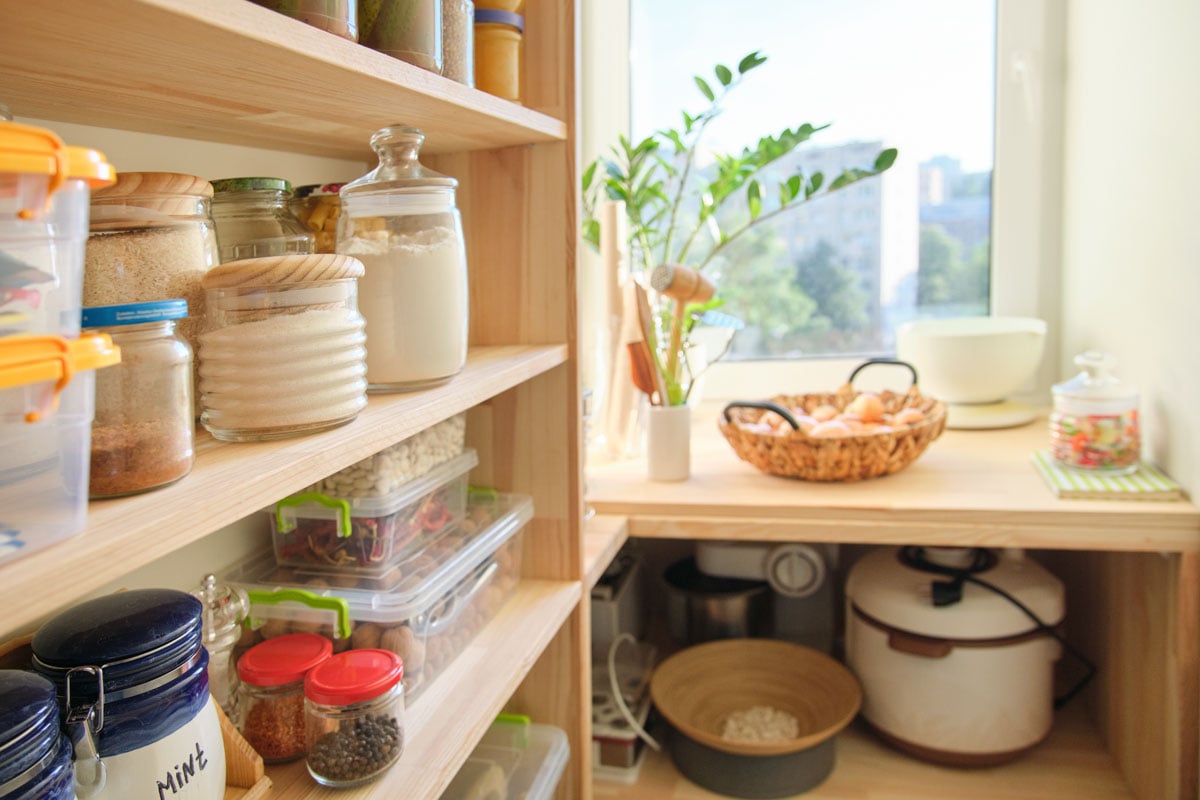 Organized pantry with shelves holding jars and containers of dry goods, a counter with kitchen appliances, a basket of onions, and a potted plant by a sunny window.