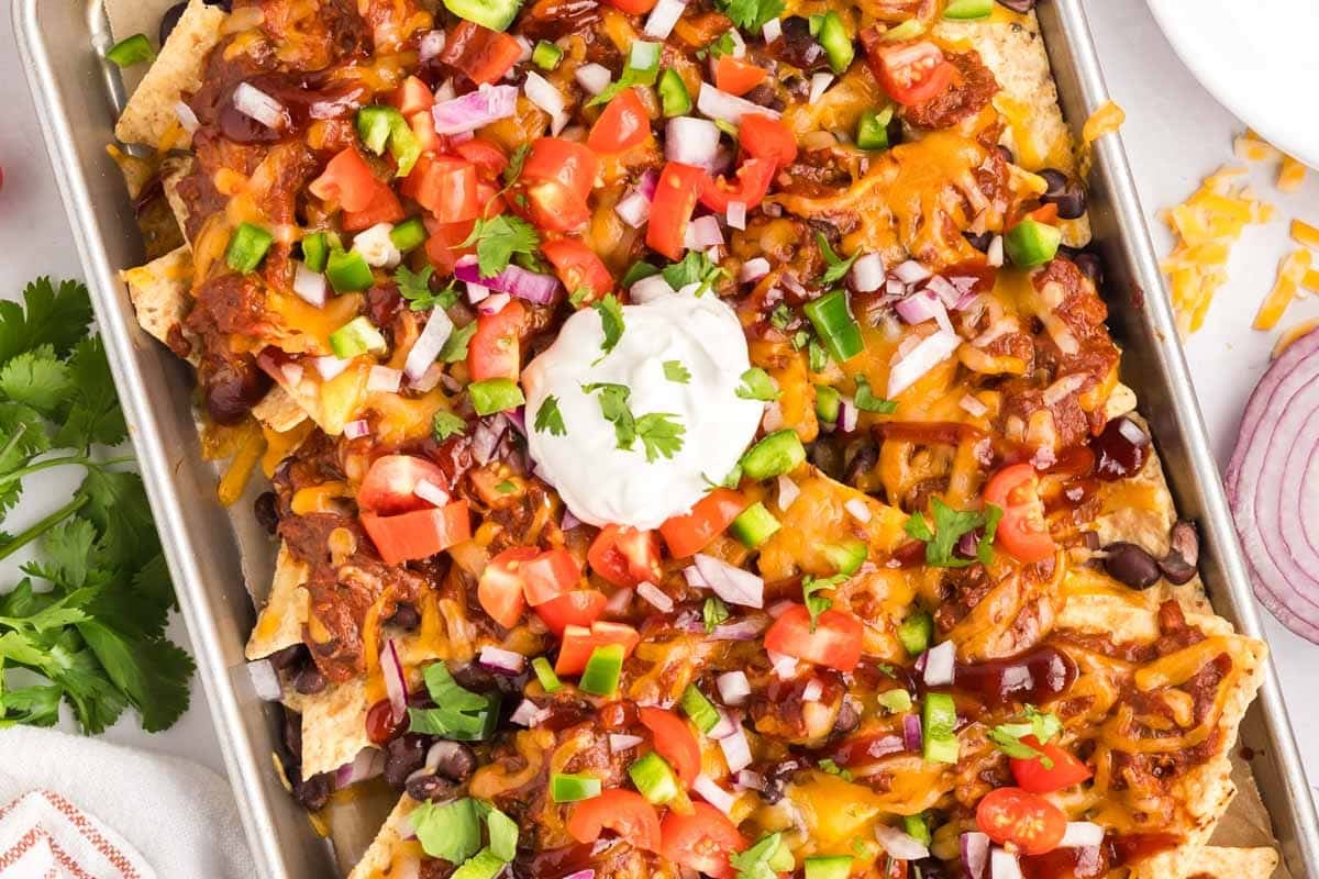 A tray of pulled pork nachos topped with melted cheese, chopped tomatoes, onions, beans, jalape&ntilde;os, and a dollop of sour cream, garnished with cilantro.