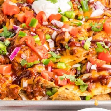 A tray of pulled pork nachos topped with melted cheese, chopped tomatoes, green and red peppers, red onions, cilantro, and a dollop of sour cream.