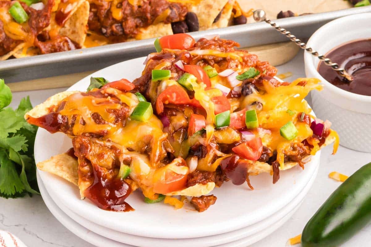 A plate of pulled pork nachos topped with melted cheese, chopped tomatoes, green onions, red onions, black beans, and barbecue sauce, with a jalape&ntilde;o and fresh cilantro nearby.