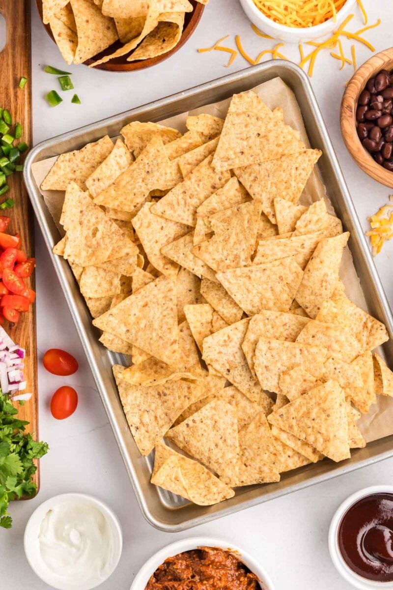 A baking sheet filled with plain tortilla chips is surrounded by small bowls of shredded cheese, black beans, tomatoes, chopped onions, sour cream, and other nacho toppings.