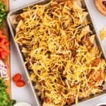 A baking sheet filled with nachos topped with shredded cheese, black beans, and chili, surrounded by bowls of fresh toppings and condiments.