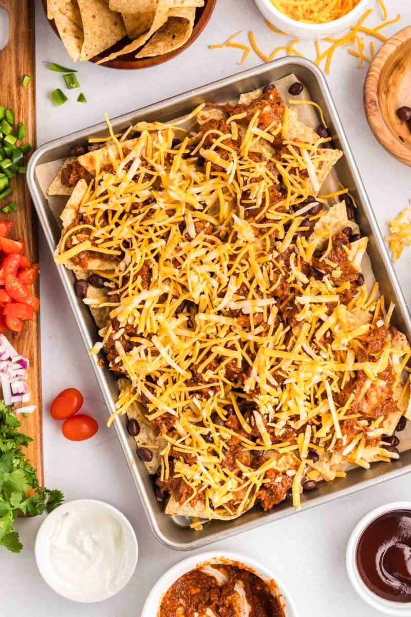 A baking sheet filled with nachos topped with shredded cheese, black beans, and chili, surrounded by bowls of fresh toppings and condiments.