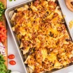 A sheet pan filled with pulled pork nachos topped with melted cheese, beans, and ground meat, surrounded by bowls of chips, toppings, sour cream, and salsa.