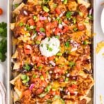 A baking sheet filled with loaded nachos topped with melted cheese, beans, chopped tomatoes, onions, jalapeños, cilantro, and a dollop of sour cream.