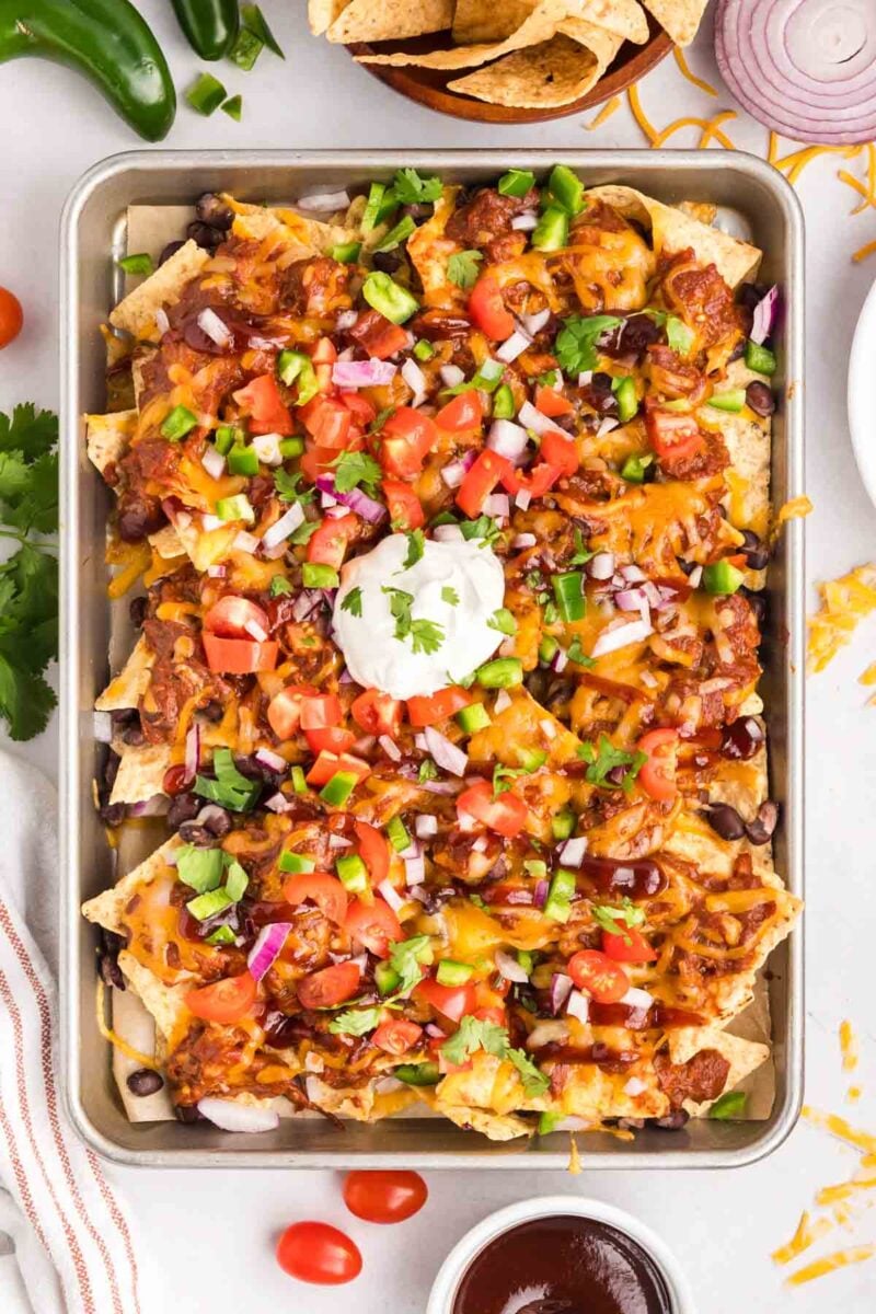 A baking sheet filled with loaded nachos topped with melted cheese, beans, chopped tomatoes, onions, jalape&ntilde;os, cilantro, and a dollop of sour cream.