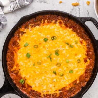 A cast iron skillet filled with cheesy baked dip topped with chopped green onions sits on a marble counter next to a striped towel and a bowl of corn chips.