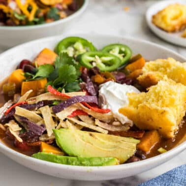A bowl of chili topped with tortilla strips, avocado slices, sour cream, jalapeños, and cilantro, served with cornbread on the side.
