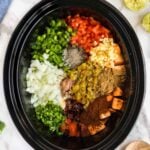 A slow cooker filled with chopped onions, bell peppers, tomatoes, green chilies, sweet potatoes, garlic, spices, and seasonings, ready to be cooked. Bowls of beans sit nearby.