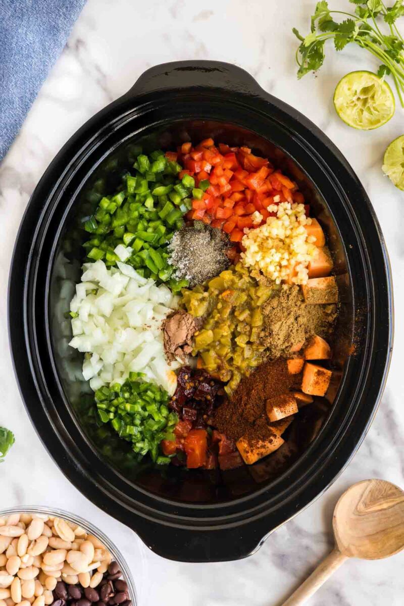 A slow cooker filled with chopped onions, bell peppers, tomatoes, green chilies, sweet potatoes, garlic, spices, and seasonings, ready to be cooked. Bowls of beans sit nearby.