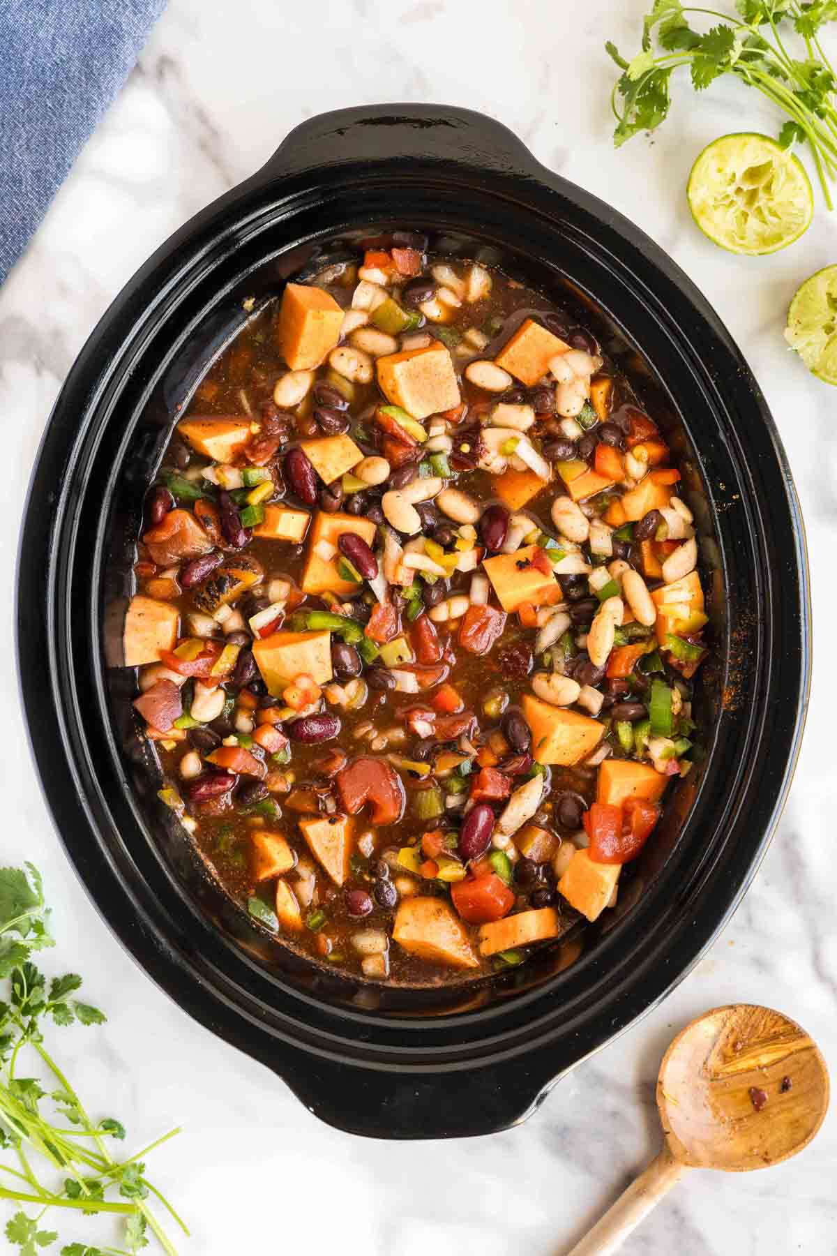 A slow cooker filled with a mixture of beans, chopped sweet potatoes, tomatoes, and vegetables in broth, surrounded by cilantro, lime halves, and a wooden spoon on a marble surface.