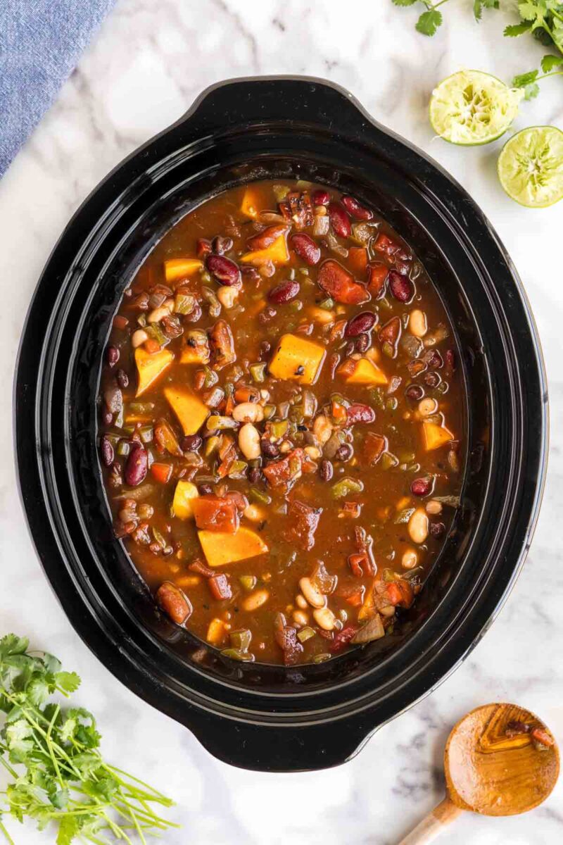 A slow cooker filled with vegetarian chili containing beans, sweet potatoes, tomatoes, and vegetables, sitting on a marble countertop with herbs and lime nearby.