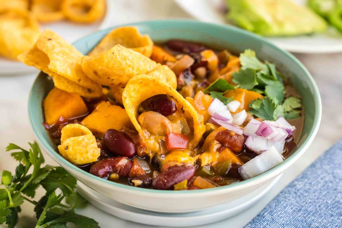 A bowl of chili topped with corn chips, chopped red onion, cilantro, and melted cheese, with beans and vegetables visible in the chili.