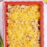A rectangular red baking dish filled with cubed ingredients topped with shredded cheddar cheese, placed on a light surface.