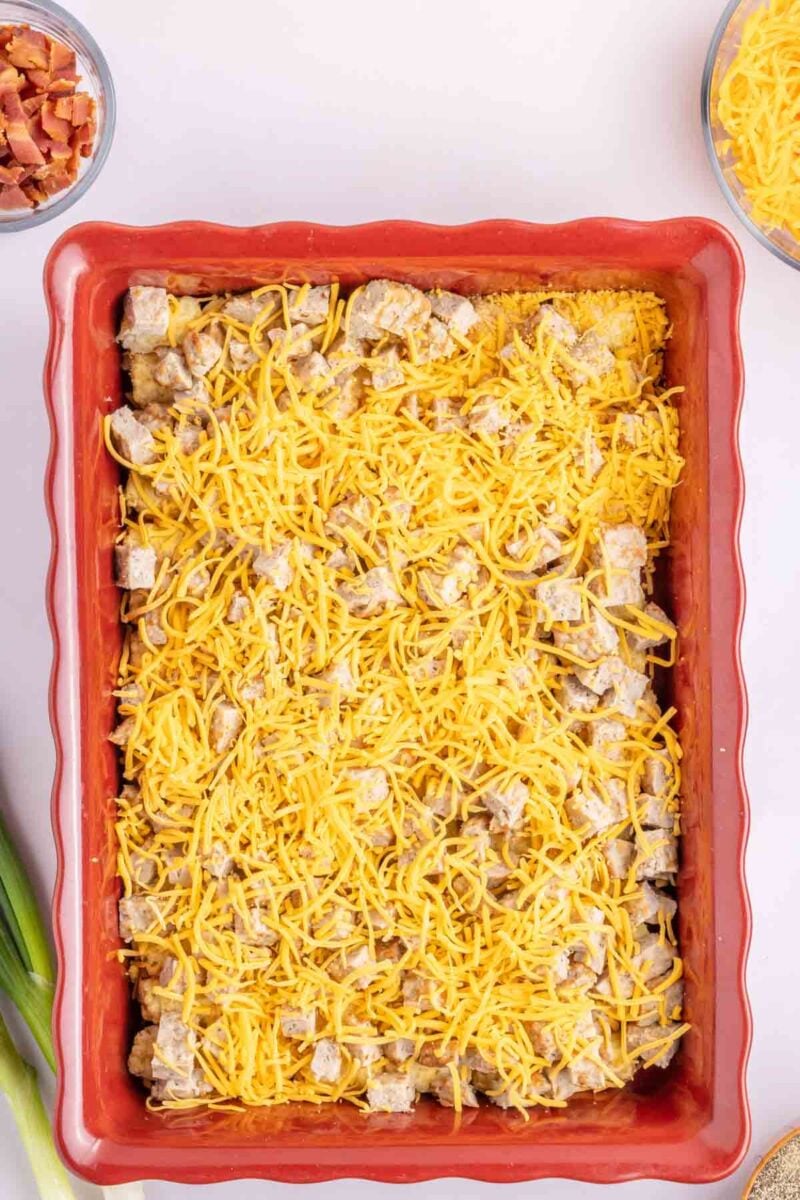 A rectangular red baking dish filled with cubed ingredients topped with shredded cheddar cheese, placed on a light surface.