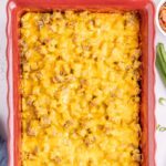 A baked tater tot breakfast casserole in a red dish topped with melted cheddar cheese, with chopped green onions and other toppings in bowls nearby.