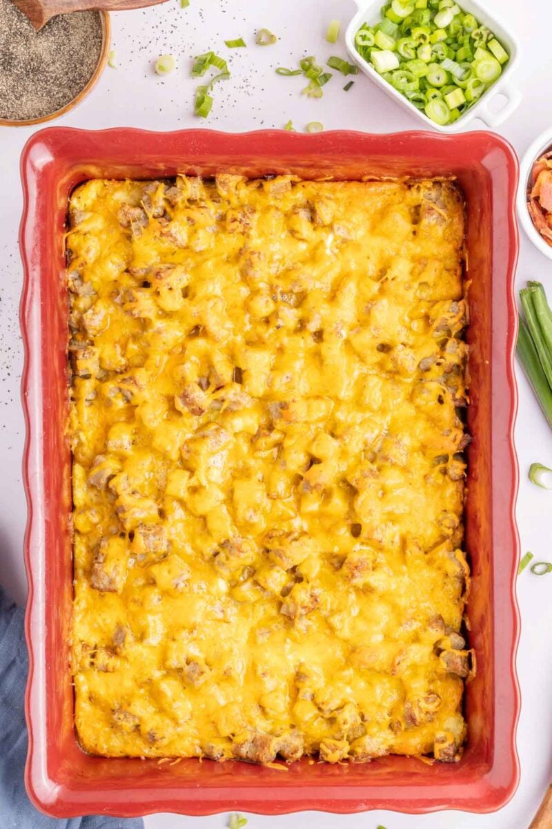A baked tater tot breakfast casserole in a red dish topped with melted cheddar cheese, with chopped green onions and other toppings in bowls nearby.