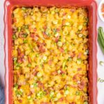 A baked casserole topped with melted cheese, chopped bacon, and sliced green onions in a red rectangular dish.