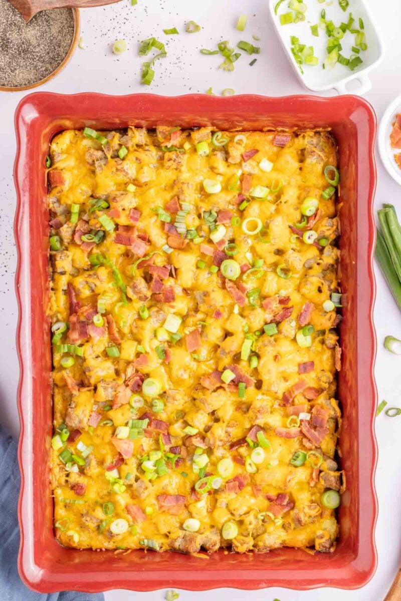 A baked casserole topped with melted cheese, chopped bacon, and sliced green onions in a red rectangular dish.